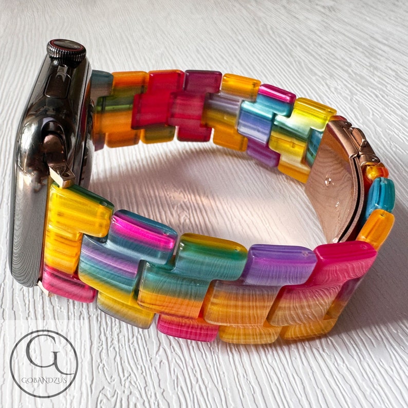 May include: A rainbow colored watch band made of plastic with a black Apple Watch attached to it.