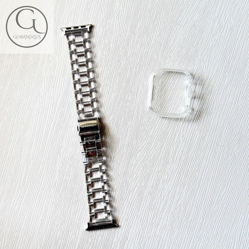 May include: A silver-toned metal link watch band featuring a rectangular clasp mechanism, accompanied by a separate clear, transparent protective bumper case designed to fit around the square housing of a digital smartwatch.