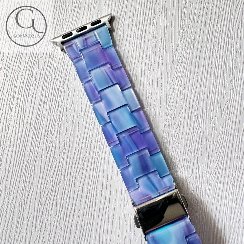 May include: A watch band with a mosaic design in shades of blue and purple. The band is made of rectangular links and has a silver-colored clasp and connectors. The band is designed to fit a smartwatch.