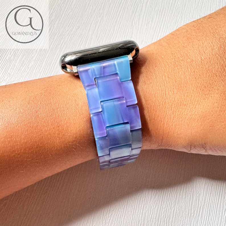 May include: A blue and purple watch band is attached to a silver watch. The band is made of rectangular links that transition from light blue to purple. The watch band is on a person's wrist. The brand name "GOBANDZUS" is in the upper left corner.