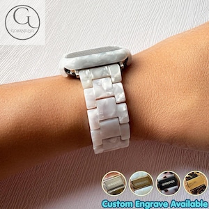 Op de afbeelding: Een wit gemarmerde horlogeband van hars met een schakelontwerp bevestigd aan een smartwatch. De band is voorzien van een gepolijste metalen sluiting. Vier kleine ronde iconen tonen verschillende kleuren sluitingen met de tekst Custom Engrave Available.