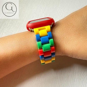 Pode incluir: Apresenta-se uma colorida pulseira de relógio em vermelho, amarelo, azul e verde. A pulseira do relógio está presa a uma caixa de relógio vermelha. A pulseira é feita de blocos retangulares de cor. O relógio está no pulso de uma pessoa.