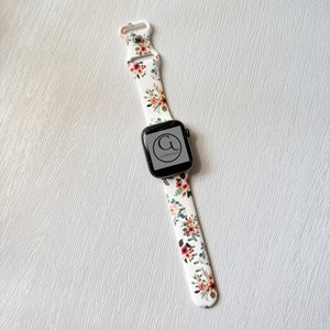 May include: White Apple Watch band with a floral print featuring pink, orange, and green flowers. The band has a black buckle and a black Apple Watch face with the text "G" and "GERMANODZA" on it.