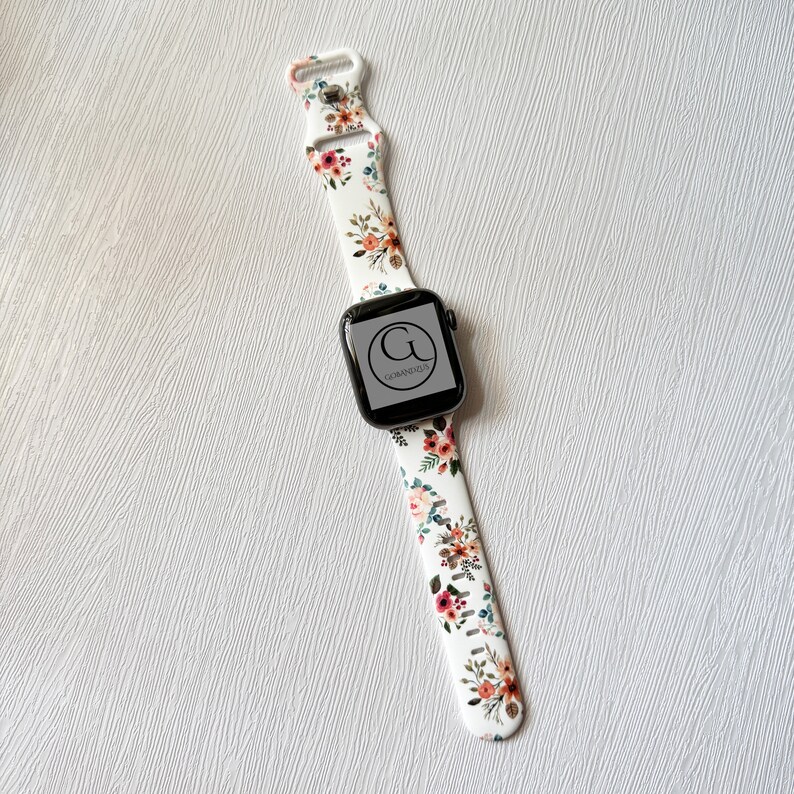 May include: White Apple Watch band with a floral print featuring pink, orange, and green flowers. The band has a black buckle and a black Apple Watch face with the text "G" and "GERMANODZA" on it.