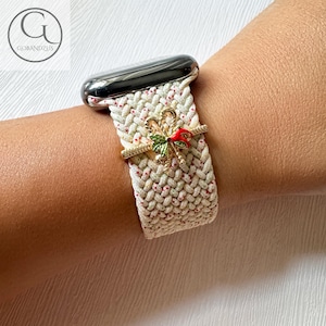 以下が含まれることがあります： 白と緑の模様のApple Watchバンドに、金と赤のリボンチャームが付いている。バンドは織り生地で作られており、繊細な花柄が施されている。