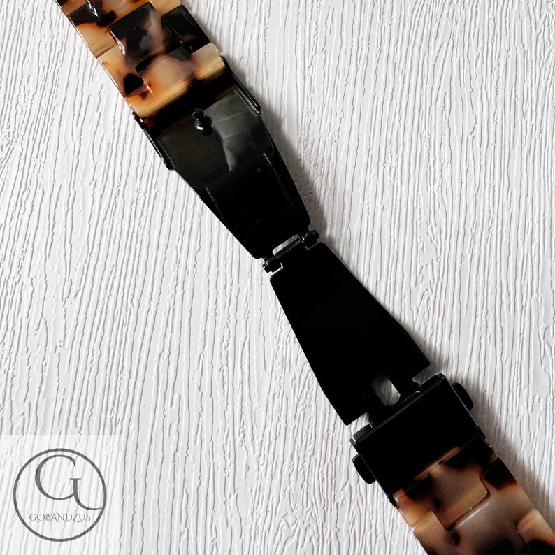 May include: A close-up of a watch band with a tortoiseshell pattern and a black metal clasp. The band is made of plastic and has a textured surface.