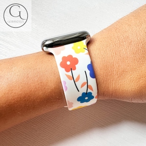 Puede incluir: Banda blanca para Apple Watch con un patrón floral colorido. La banda presenta flores rosas, azules, amarillas y naranjas con tallos y hojas negras.