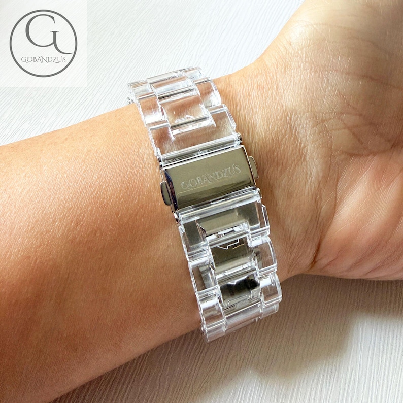 May include: A clear, transparent watch band featuring interlocking links made of a smooth, hard material. The band includes a silver-toned metal deployment clasp with the brand name GOBANDZUS engraved on the surface, securing the links together.