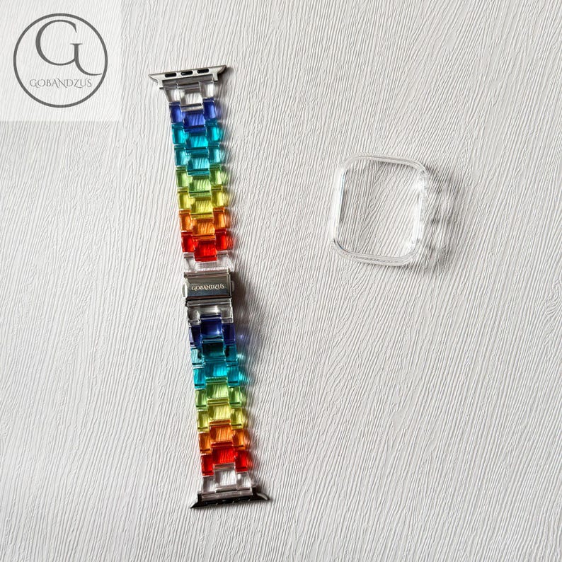 May include: A rainbow-colored link watch band made of translucent resin segments with a silver-toned metal clasp featuring the text GOBANDZUS, accompanied by a separate clear, rectangular protective watch case cover for a digital timepiece.
