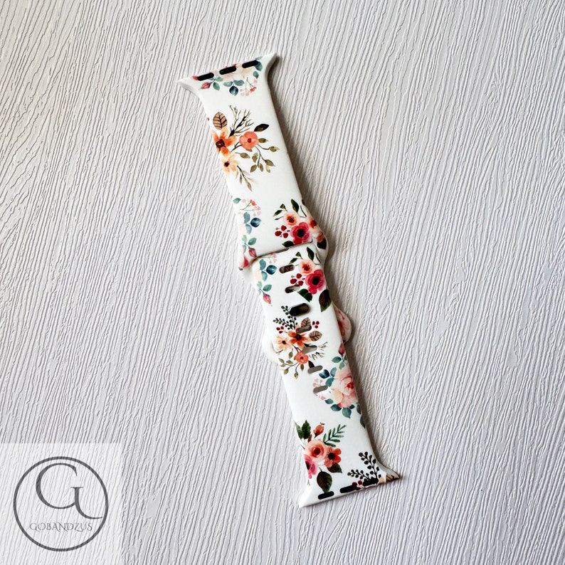 May include: White silicone Apple Watch band with a floral print featuring pink, red, and orange flowers. The band has a white background with green leaves.