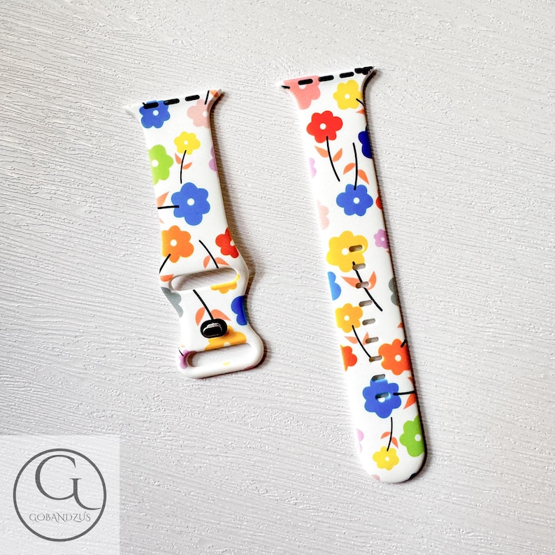 May include: A white Apple Watch band with a colorful floral pattern. The band has a white background with blue, yellow, orange, red, and green flowers. The band is made of silicone and is compatible with Apple Watch Series 4 and later.