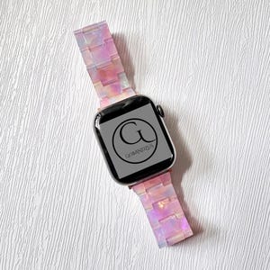 May include: A pink and white iridescent Apple Watch band with a black watch face that says "GOBANDZUS".
