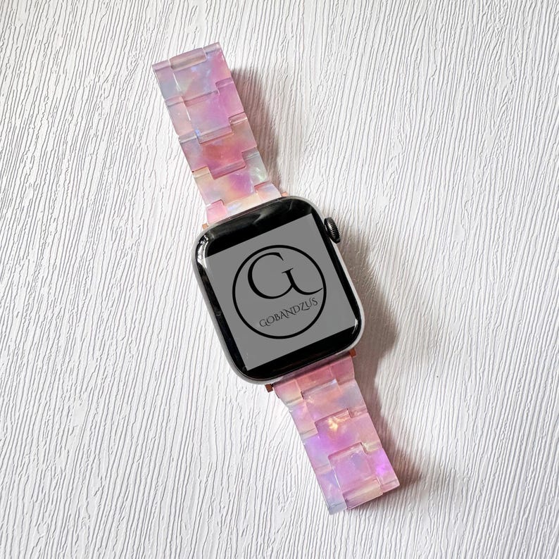 May include: A pink and white iridescent Apple Watch band with a black watch face that says "GOBANDZUS".