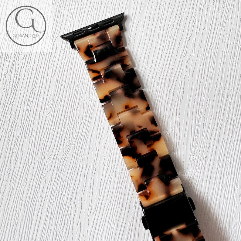 May include: A tortoiseshell patterned Apple Watch band with a black metal clasp.