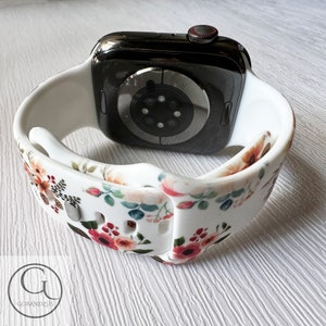 May include: White silicone Apple Watch band with a floral print. The band has a white background with red, pink, and green flowers. The band is shown on a black Apple Watch.