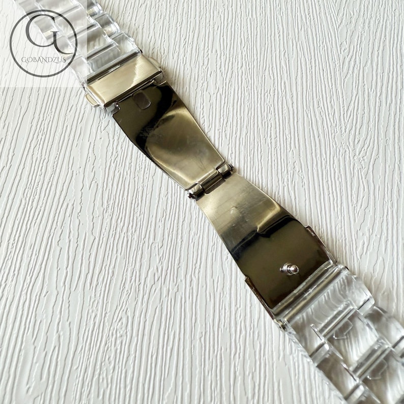 May include: A clear, transparent watch band featuring interlocking links and a polished silver-tone metal deployment clasp. The band includes a logo mark with the text GOBANDZUS. The design consists of a series of rectangular, translucent segments.