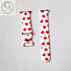 May include: White Apple Watch band with a red heart pattern. The band is made of silicone and has a buckle closure.