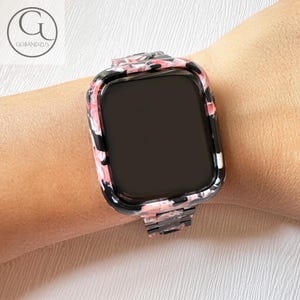 Puede incluir: Un reloj inteligente con una carcasa protectora y una correa de eslabones con un patr&oacute;n jaspeado en tonos negro, blanco y rosa suave. El reloj tiene una pantalla negra rectangular y un acabado liso y brillante en el marco y la correa.