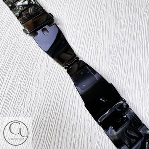 May include: Black and white patterned watch band with a black metal clasp. The band is made of a hard plastic material.