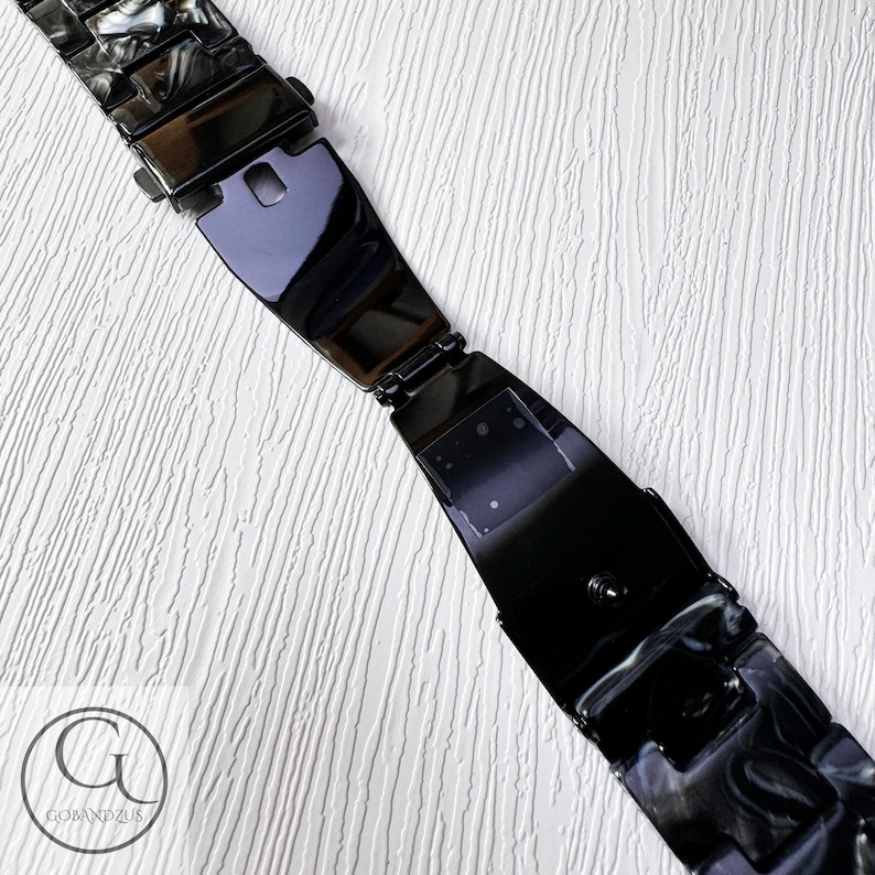 May include: Black and white patterned watch band with a black metal clasp. The band is made of a hard plastic material.