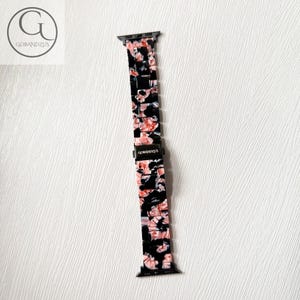 Puede incluir: Una correa de reloj con un patr&oacute;n de m&aacute;rmol en negro, blanco y rosa con eslabones rectangulares. La correa incluye un cierre de metal negro grabado con el texto GOBANDZUS y conectores de metal negro en ambos extremos para fijar al reloj.