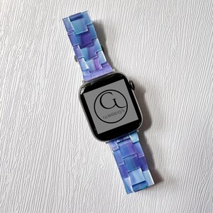 May include: A smartwatch with a blue and purple band. The band has a mosaic-like design. The watch face displays the logo "G BANDZUS" in a circular design. The watch is set against a white textured background.