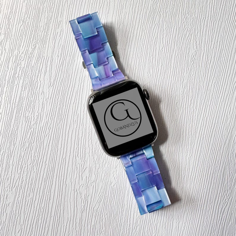 May include: A smartwatch with a blue and purple band. The band has a mosaic-like design. The watch face displays the logo "G BANDZUS" in a circular design. The watch is set against a white textured background.