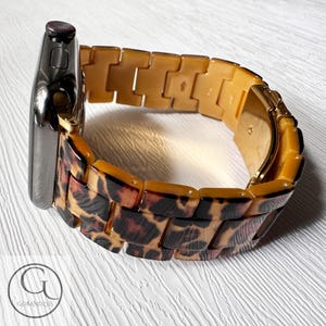 Puede incluir: Una correa de reloj de pl&aacute;stico para Apple Watch con estampado de leopardo dorado y marr&oacute;n. La correa es de estilo de eslabones.