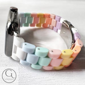 Op de afbeelding: Een pastelkleurig regenboogkleurig Apple Watch-bandje met een witte basis en zilveren accenten. Het bandje is gemaakt van plastic en heeft een uniek, kleurrijk ontwerp.