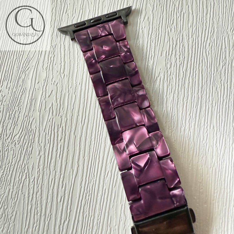 May include: A watch band featuring a purple marbled resin link design with a dark metal connector at the top. The links have a glossy finish with varying shades of deep violet and lighter purple tones, creating a textured appearance throughout.