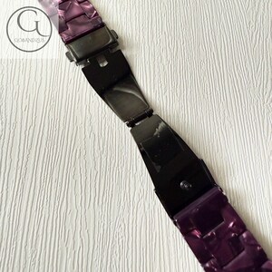 May include: A purple watch band featuring a glossy, marbled finish with a black metal deployment clasp in the center. The band consists of rectangular links and includes a circular logo with the text GOBANDZUS in the upper left corner.