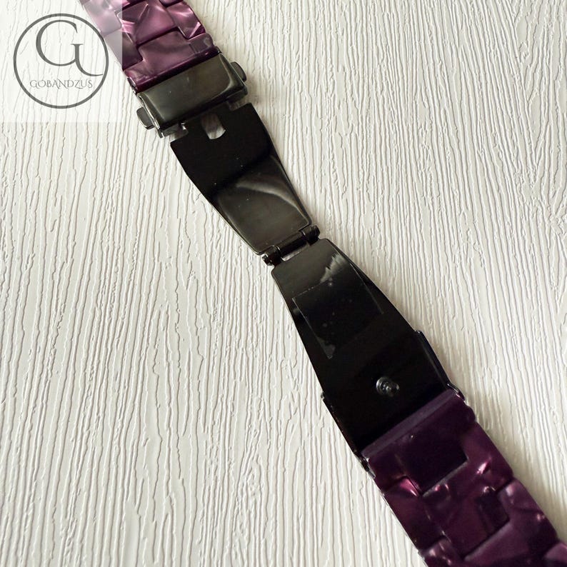 May include: A purple watch band featuring a glossy, marbled finish with a black metal deployment clasp in the center. The band consists of rectangular links and includes a circular logo with the text GOBANDZUS in the upper left corner.