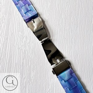 May include: A blue and purple bracelet with a woven design. The bracelet has a silver clasp and is displayed on a white textured surface. The bracelet is made of rectangular pieces of blue and purple material.