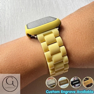 Puede incluir: Una caja de reloj inteligente de color amarillo pálido con una correa de eslabones a juego. La correa consta de segmentos rectangulares entrelazados. Cuatro miniaturas circulares abajo muestran cierres metálicos con el texto Custom Engrave Available.