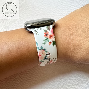 May include: White Apple Watch band with a floral print featuring pink, red, and green flowers. The band is made of silicone and has a black metal buckle.