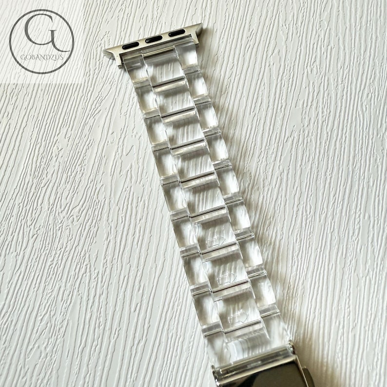 May include: A clear, transparent watch band featuring a linked, multi-segment design with a metallic silver-toned connector at the top. The band is constructed from smooth, rigid material and includes a secure metal clasp mechanism at the bottom end.