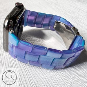 May include: A close-up of a smartwatch with a blue and purple link band. The band is made of rectangular links that transition from light blue to purple. The watch case is black with a red button.