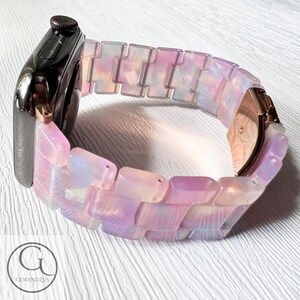 May include: A pink and white iridescent watch band with a rose gold clasp. The band is made of a series of rectangular links.