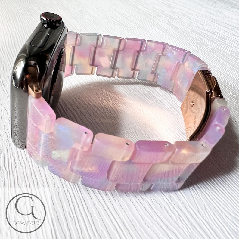 May include: A pink and white iridescent watch band with a rose gold clasp. The band is made of a series of rectangular links.
