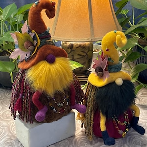 May include: Two whimsical gnome figures. One gnome has a rust-colored hat with a yellow beard and purple feet. The other gnome has a yellow hat and a black beard. Both gnomes have beaded necklaces and are decorated with flowers and autumn leaves.