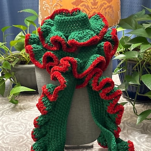 May include: A handmade green scarf with red trim and ruffled edges. The scarf is made of a soft, textured yarn and is displayed on a gray mannequin head. The scarf's design features a vibrant green color with contrasting red accents.