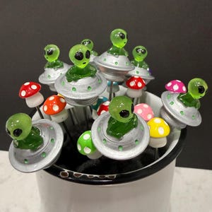 May include: A collection of whimsical alien-themed decorative picks. Each pick features a green alien figure atop a silver UFO or a colorful mushroom. The picks are arranged in a white enamel mug, creating a playful and unique display.