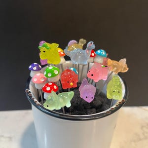May include: A white ceramic mug filled with colorful dinosaur and mushroom shaped food picks. The picks are made of glass and have a shiny finish.