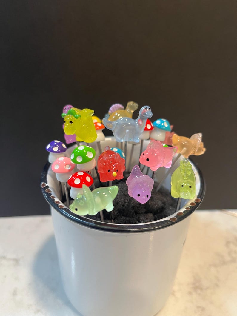 May include: A white ceramic mug filled with colorful dinosaur and mushroom shaped food picks. The picks are made of glass and have a shiny finish.