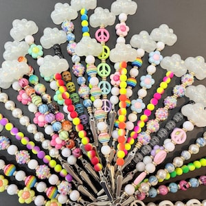 May include: A collection of colorful beaded phone charms with various charms, including clouds, peace signs, flowers, and skulls. The charms are attached to silver clips.