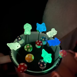May include: A collection of glow-in-the-dark dinosaur figurines and mushroom-shaped decorations in a black cup. The dinosaurs are green, blue, and white. The mushrooms are red and white with black polka dots.