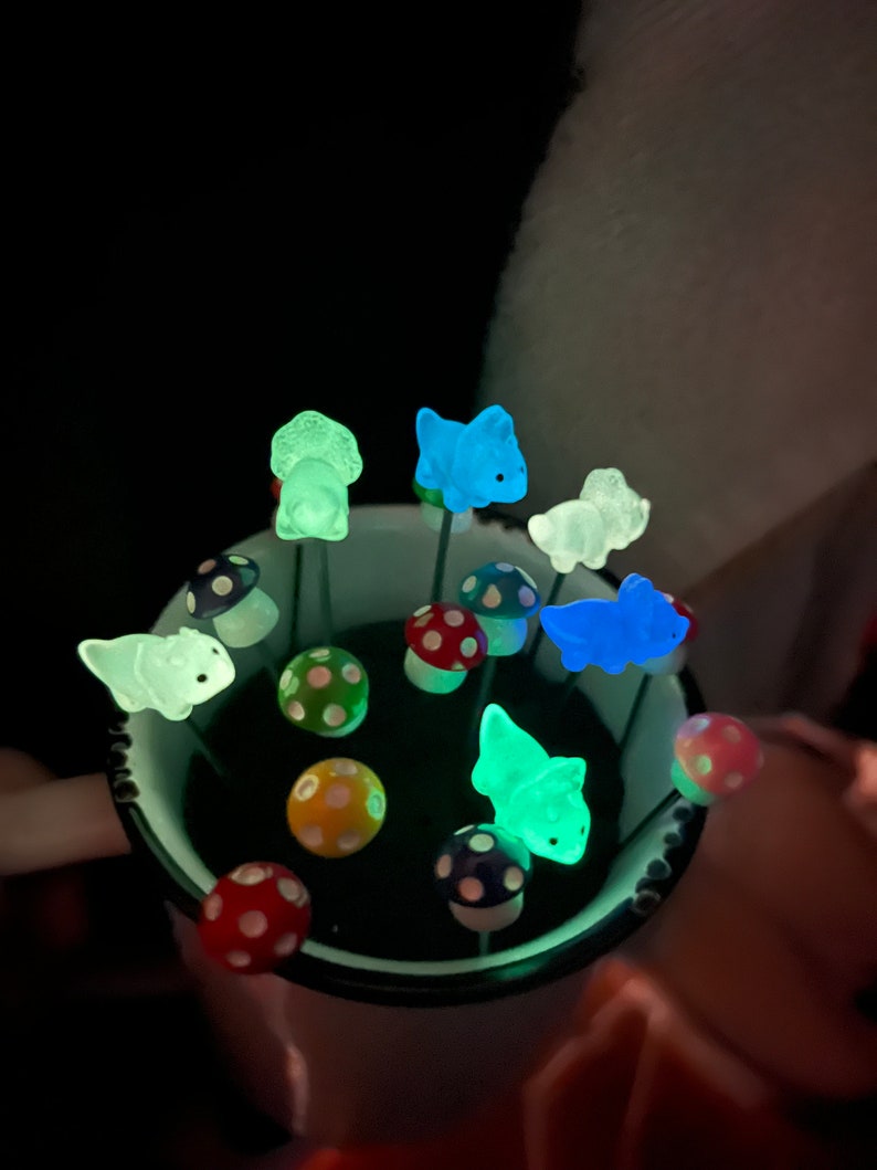 May include: A collection of glow-in-the-dark dinosaur figurines and mushroom-shaped decorations in a black cup. The dinosaurs are green, blue, and white. The mushrooms are red and white with black polka dots.