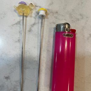 May include: Two metal skewers with small plastic figurines on top. The figurines are a yellow dinosaur and a yellow mushroom. A pink lighter is in the background.