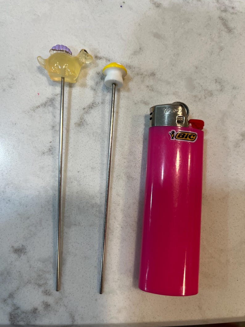 May include: Two metal skewers with small plastic figurines on top. The figurines are a yellow dinosaur and a yellow mushroom. A pink lighter is in the background.