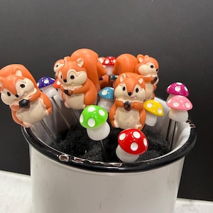 May include: A collection of decorative skewers featuring orange squirrels and colorful mushroom designs. The skewers are arranged in a white metal container with a black rim, set against a dark background.